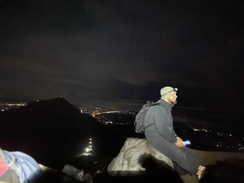 Man Sitting On Rock Nighttime City Lights Headlamp Backpack